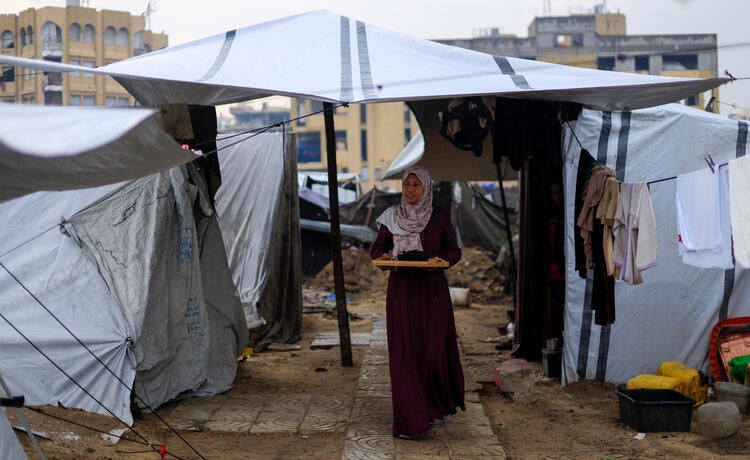 A woman in a long maroon dress and pale flowery head scarf carries a wooden tray as she walks between makeshift tents surrounded by muddy ground.